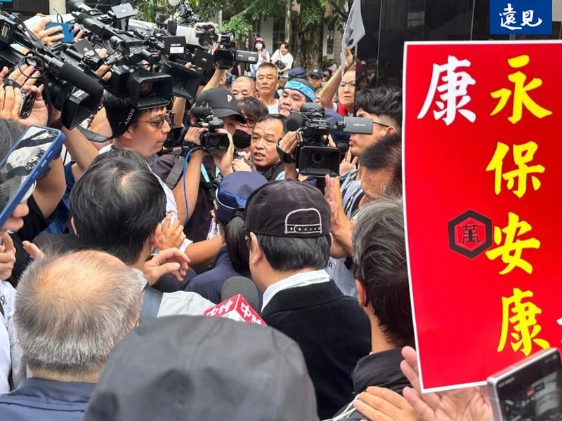 蔡康永戴黑帽在蔣萬安競總被攝影記者包圍，旁邊有人舉著寫有康永保安康的紅色牌子