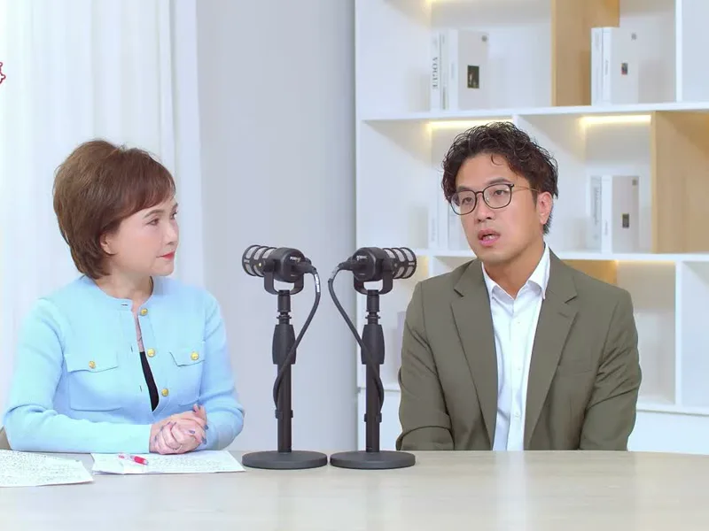 沈伯洋穿橄欖綠西裝坐在麥克風後回答問題，沈春華雙手交握在旁邊聽